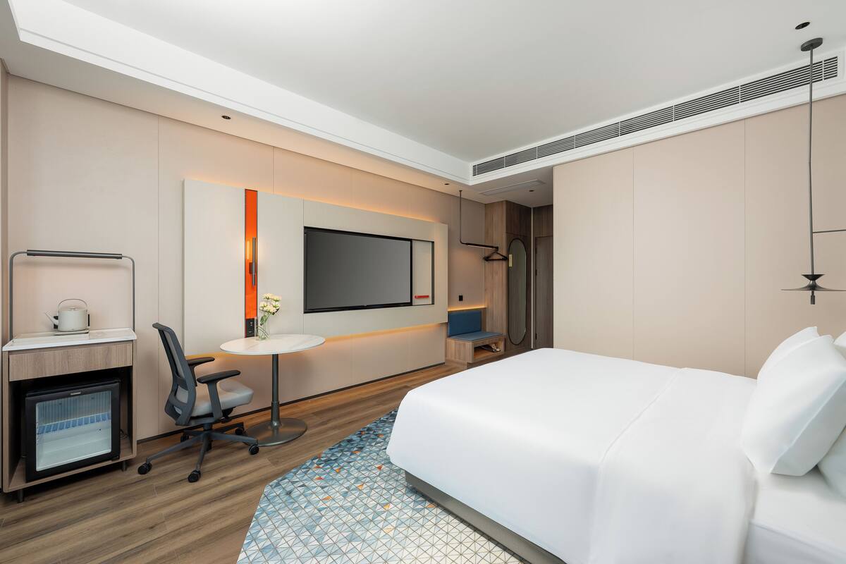 A modern hotel room with a bed, desk, chair, TV, and a small kitchenette.
