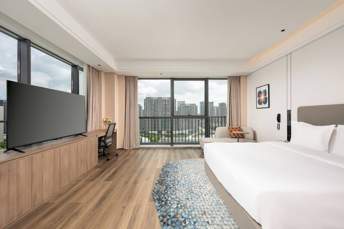 A modern hotel room with a large bed, a desk with a flat-screen TV, a chair, and a view of the city through large windows.