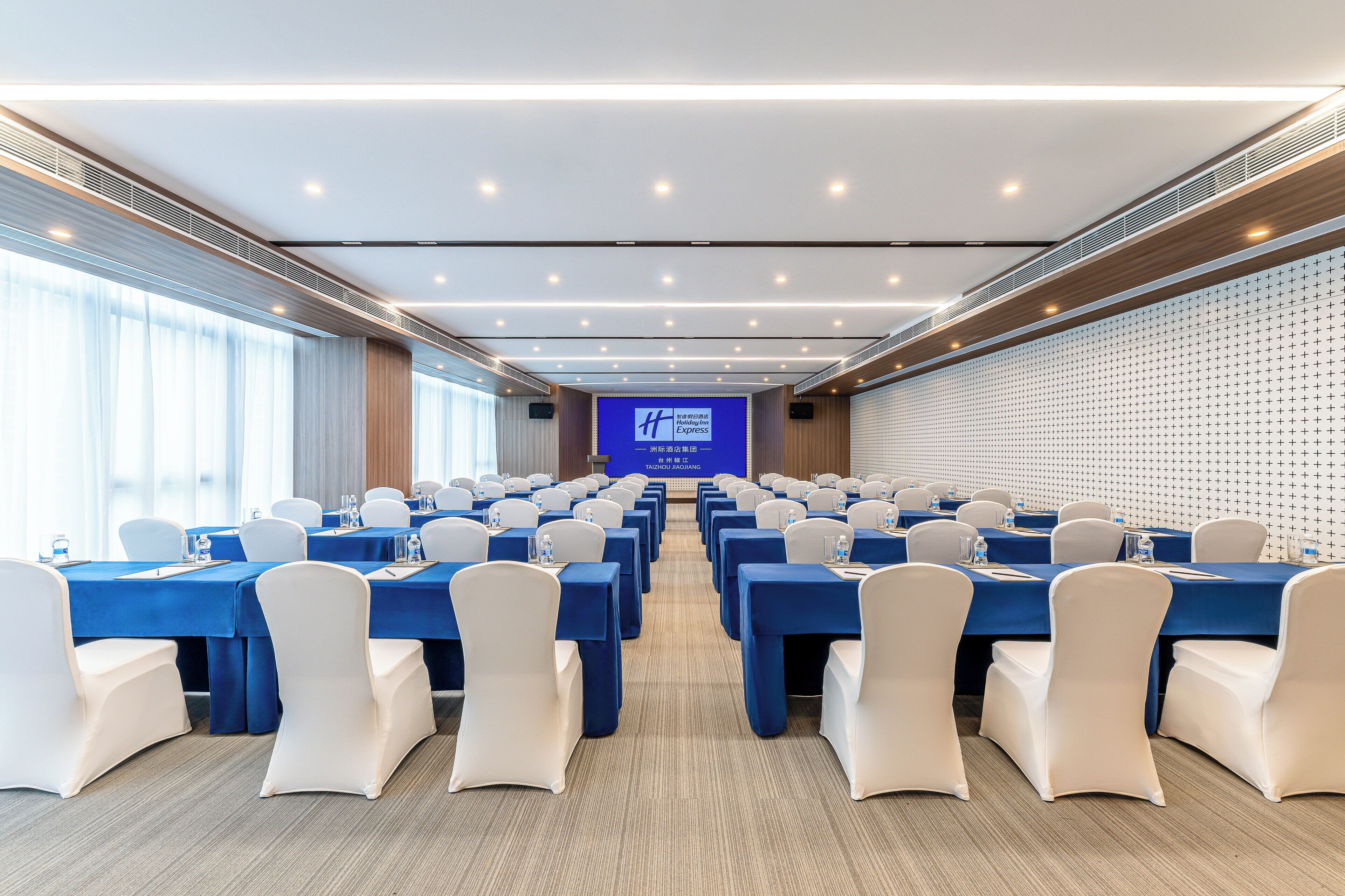Une salle de conférence avec des rangées de tables, des chaises et un grand écran à l’avant.