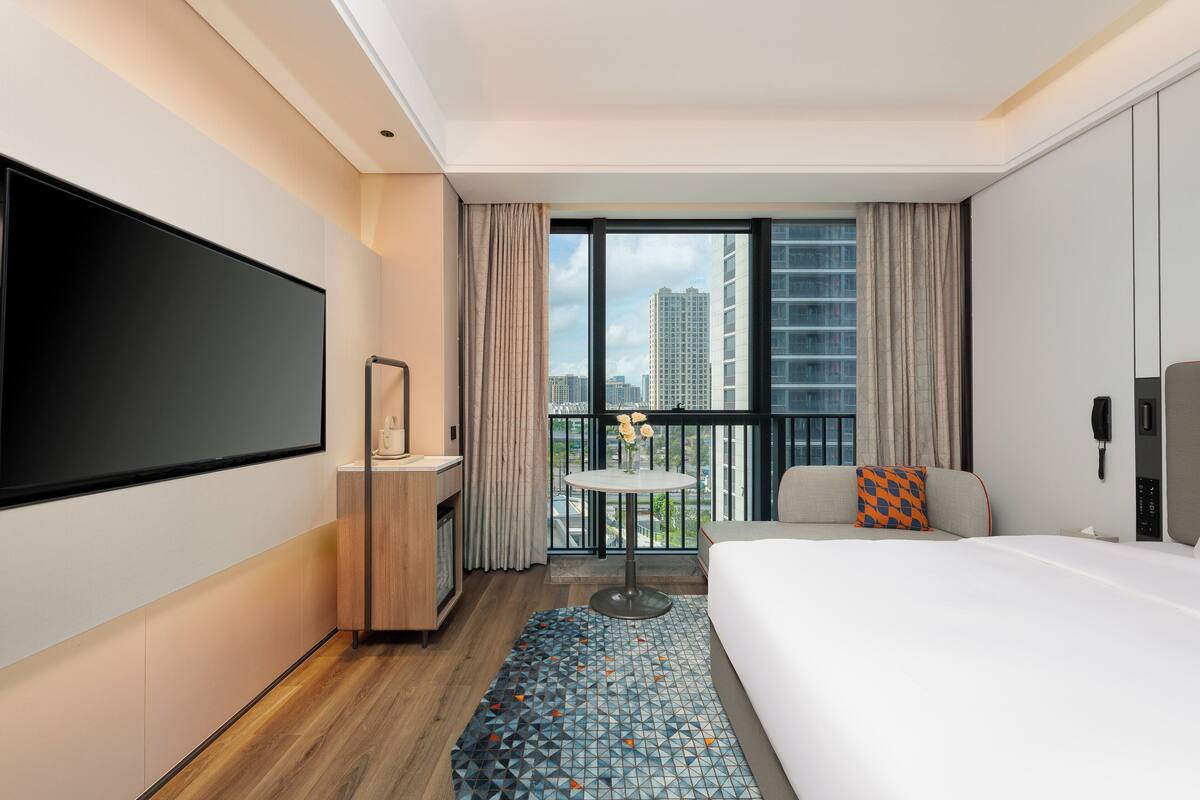 A hotel room with a large bed, a TV mounted on the wall, a small table with a vase, and a balcony with a view of high-rise buildings.