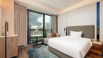 A hotel room with a large bed, a small seating area, a balcony with curtains, and a view of the city.