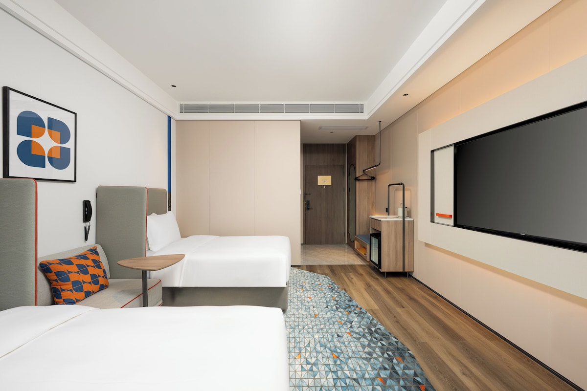 Habitación de hotel moderna con dos camas, un televisor, un escritorio y un baño visible a través de una puerta abierta.