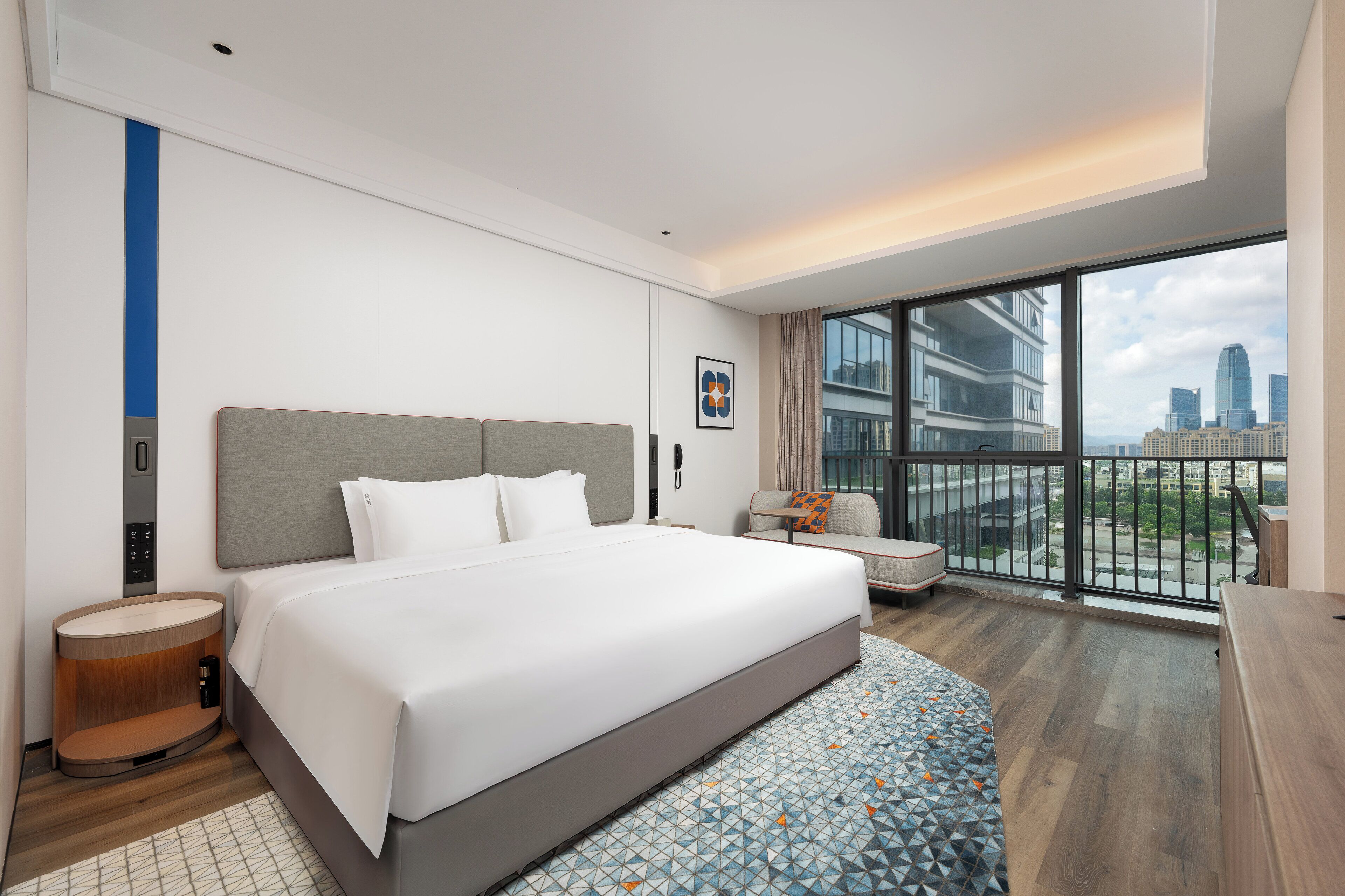 Ein modernes Hotelzimmer mit einem großen Bett, einer kleinen Sitzecke, einem Schreibtisch und einem Balkon mit Blick auf die Stadt.
