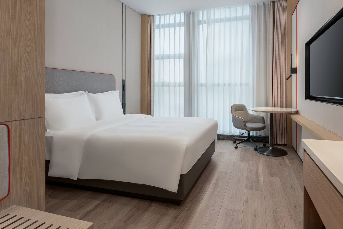 Une chambre d’hôtel avec un lit, un bureau, une chaise, une télévision et une fenêtre avec des rideaux.