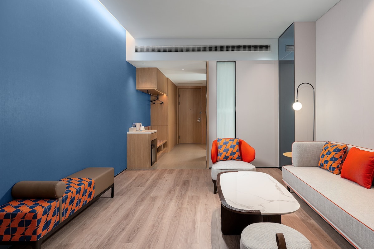 Une chambre d’hôtel moderne avec un mur d’accent bleu, un canapé agrémenté de coussins à motifs orange et bleu, une petite table et un banc avec un coussin à motifs géométriques.