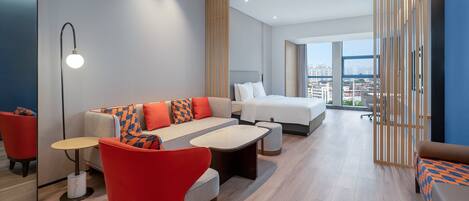 A modern hotel room with a sofa, a bed, a desk, and a view of the city.