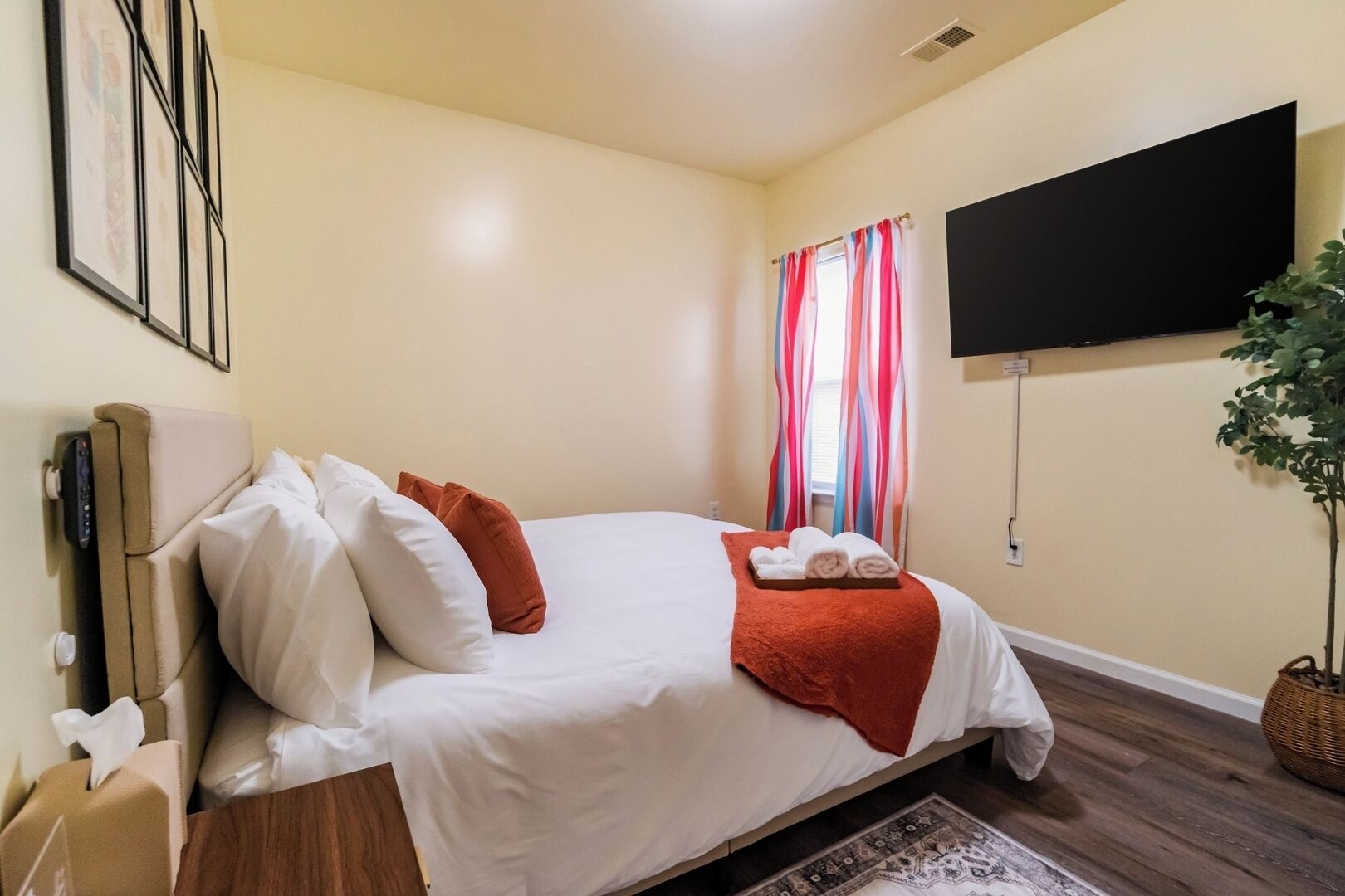 A neatly made bed with white and orange pillows, a red throw blanket, and two rolled towels on a wooden floor with a patterned rug.