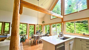 A spacious kitchen with large windows, wooden beams, and a dining area with a view of greenery. - Cedar & Salt – Forest Retreat by the Beach with Hot Tub, Sauna & EV Charger (Tofino)