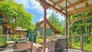 A wooden deck with outdoor furniture, a covered area, and a view of a green house with a window and a door. - Cedar & Salt – Forest Retreat by the Beach with Hot Tub, Sauna & EV Charger (Tofino)