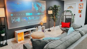 Un salon moderne doté d’un grand téléviseur à écran plat affichant l’interface d’un service de diffusion en continu, d’un confortable canapé gris, d’un fauteuil suspendu et d’une petite table d’appoint avec une lampe.