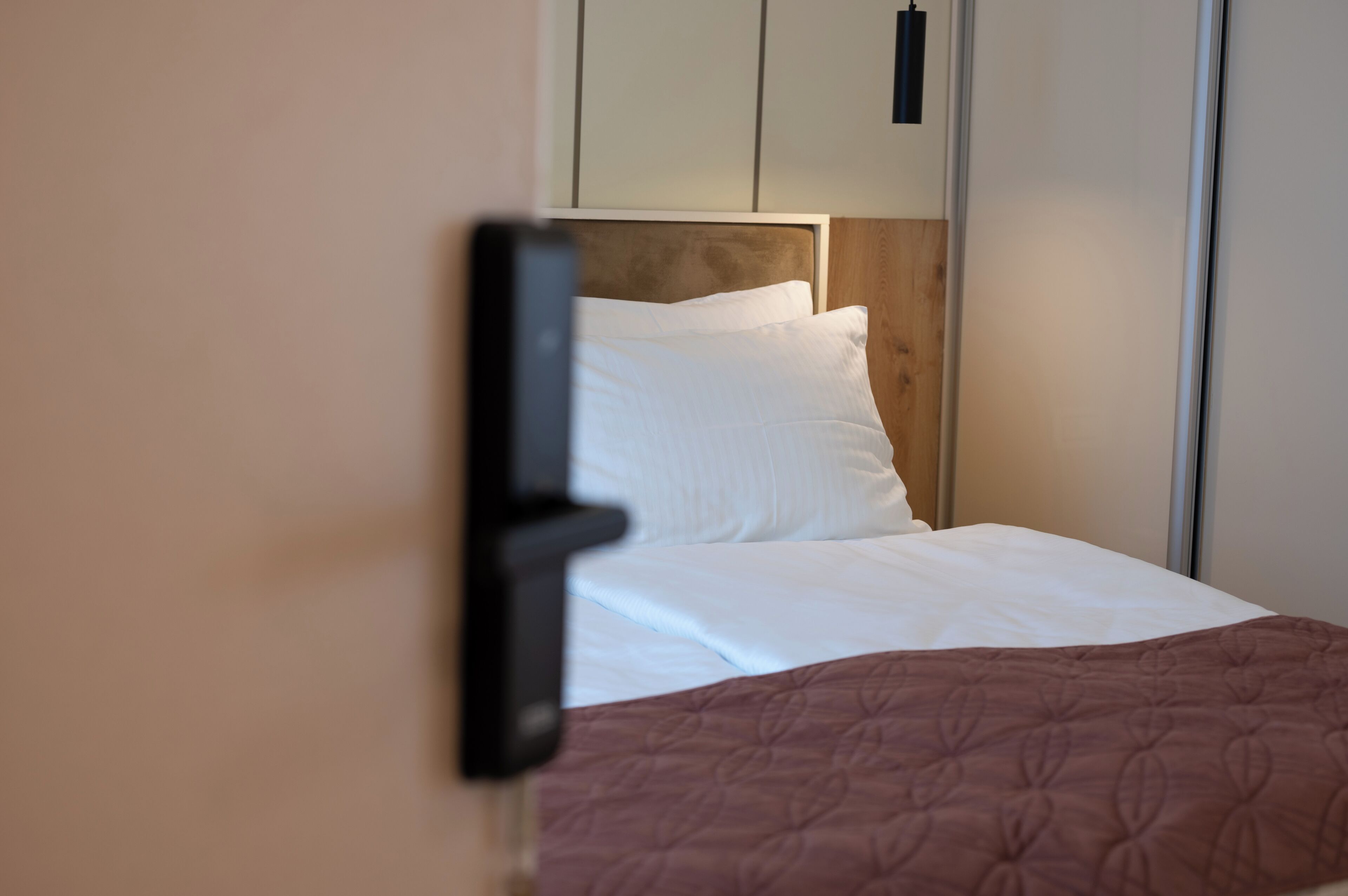 A neatly made bed with white linens and a brown quilt, a wooden headboard, and a white wardrobe with a black handle.