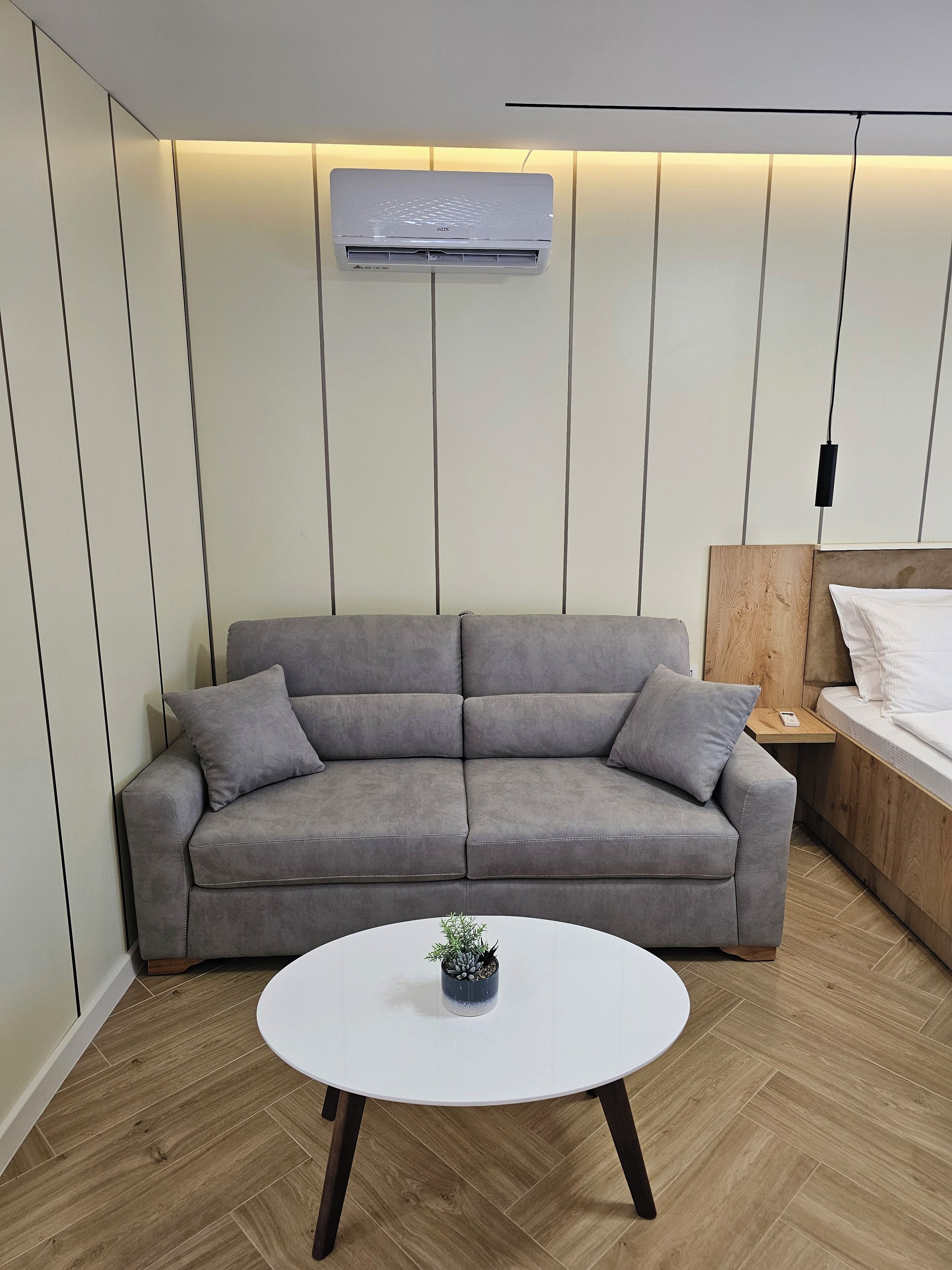 A modern living room with a grey sofa, a round white table with a plant, and a wooden headboard bed.