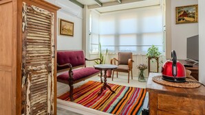 Ein gemütliches Wohnzimmer mit Sofa, Sessel und einem kleinen Tisch. Ein farbenfroher Streifen Teppich, ein roter Wasserkocher und ein Bild an der Wand sind zu sehen.