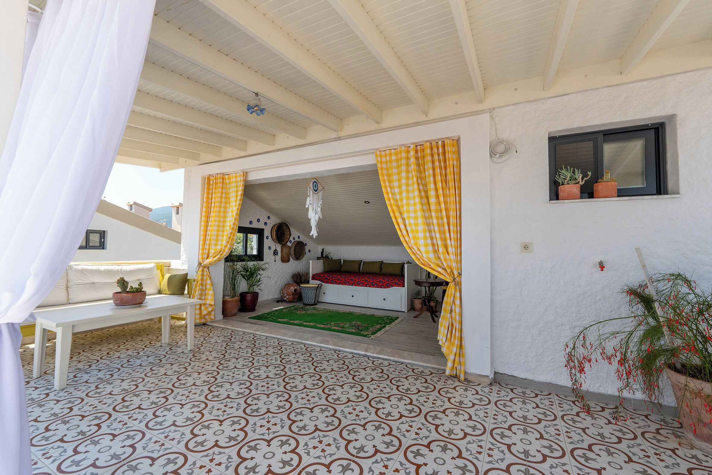 A covered outdoor area with a patterned floor, a white bench, a sofa, and a bed with a red and black patterned bedspread.