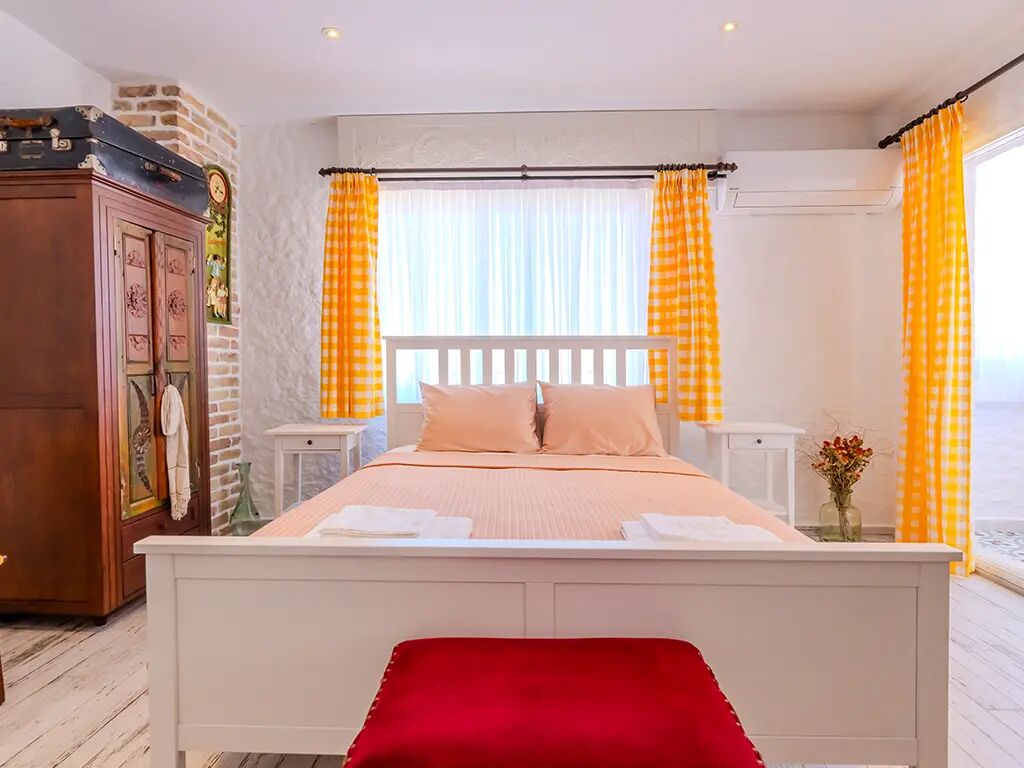 A bedroom with a bed, a red bench, a wooden wardrobe, and a window with curtains.