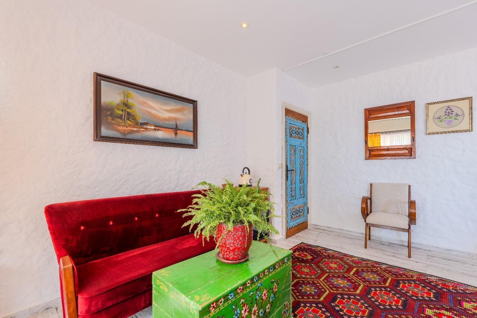 A room with a red velvet sofa, a green box with a plant, a patterned rug, and a blue door.