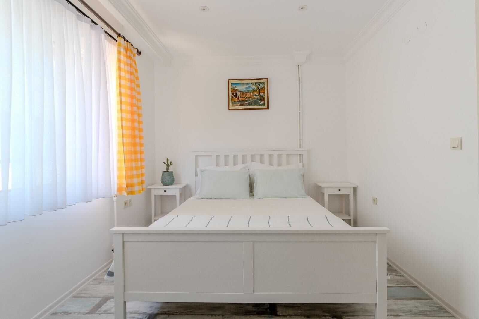 A white bedroom with a bed, two bedside tables, a window with white curtains, and a painting on the wall.