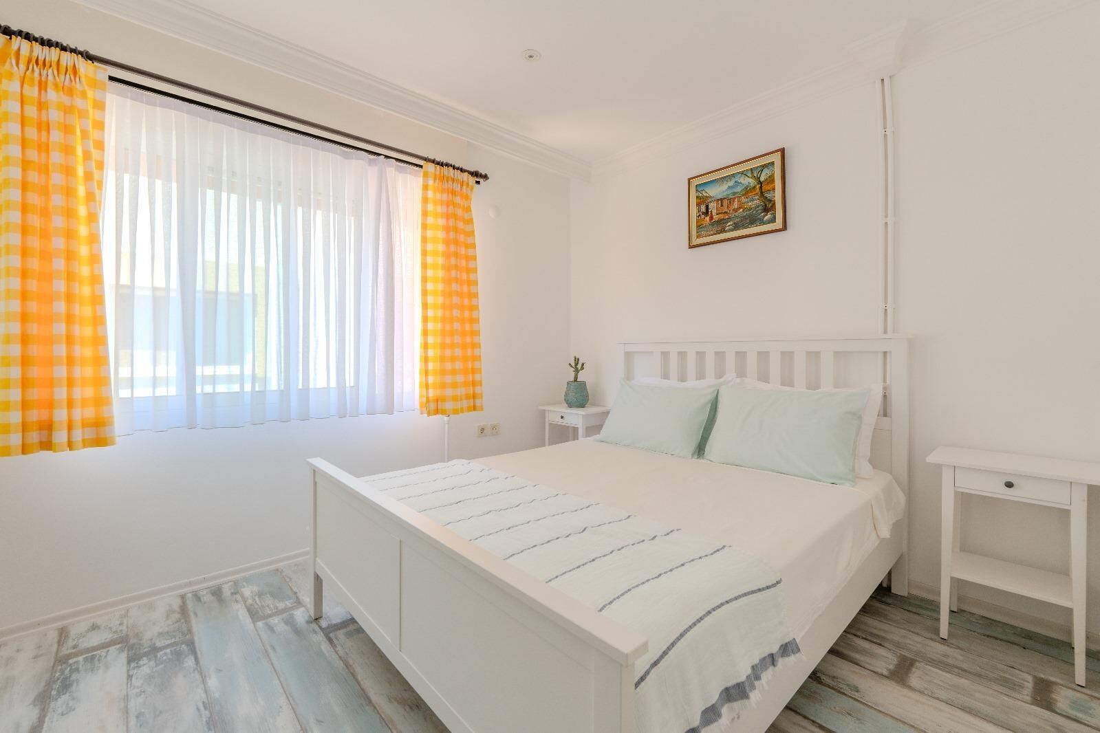 A bedroom with a white bed, a nightstand, a small table, and a window with yellow curtains.