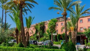 A hotel with a row of palm trees in the foreground, a parking lot with several cars, and a building with a red facade and multiple windows.