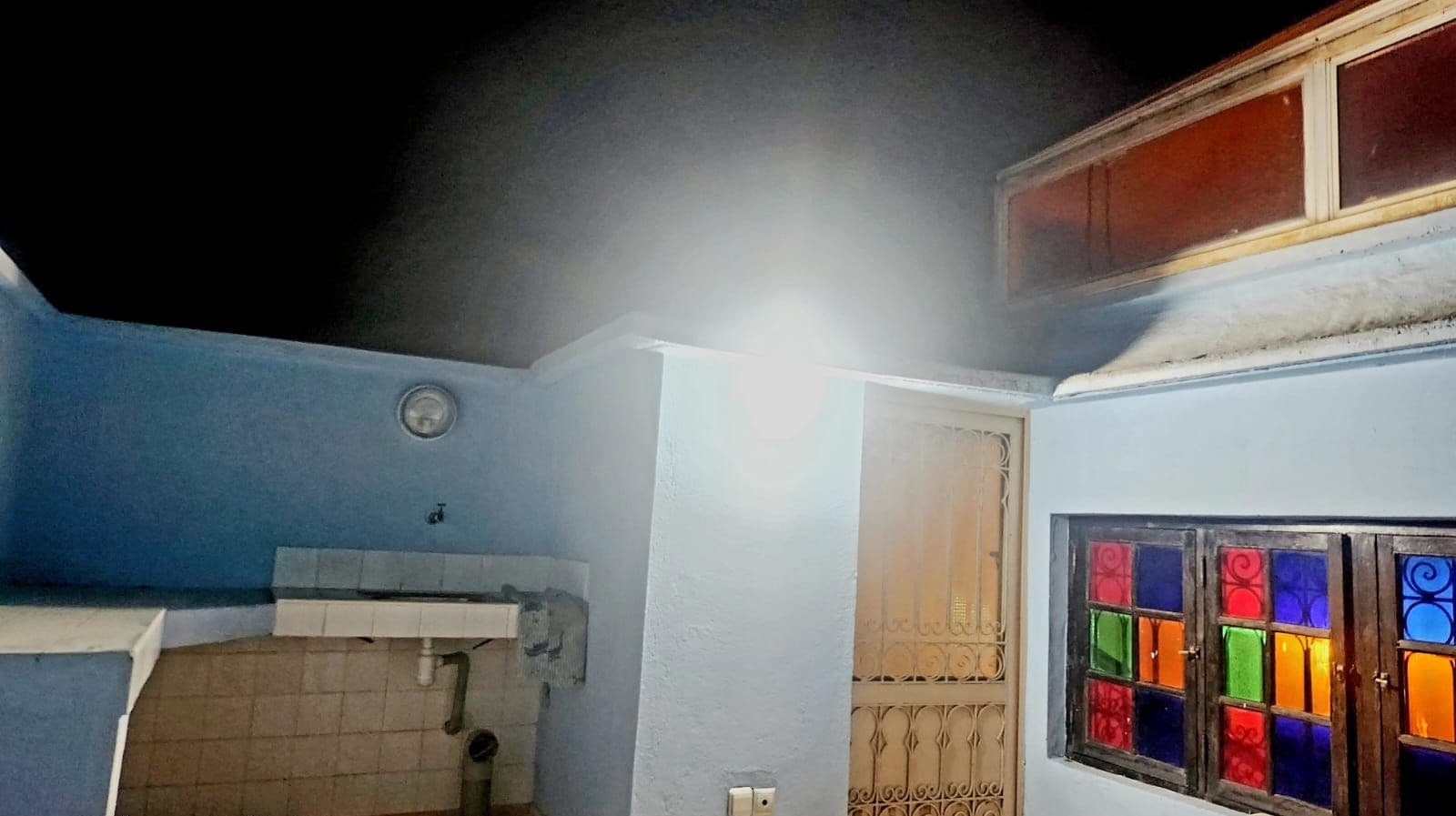 A small, dimly lit room with a sink, a door with decorative metalwork, and a window with colorful glass panes.