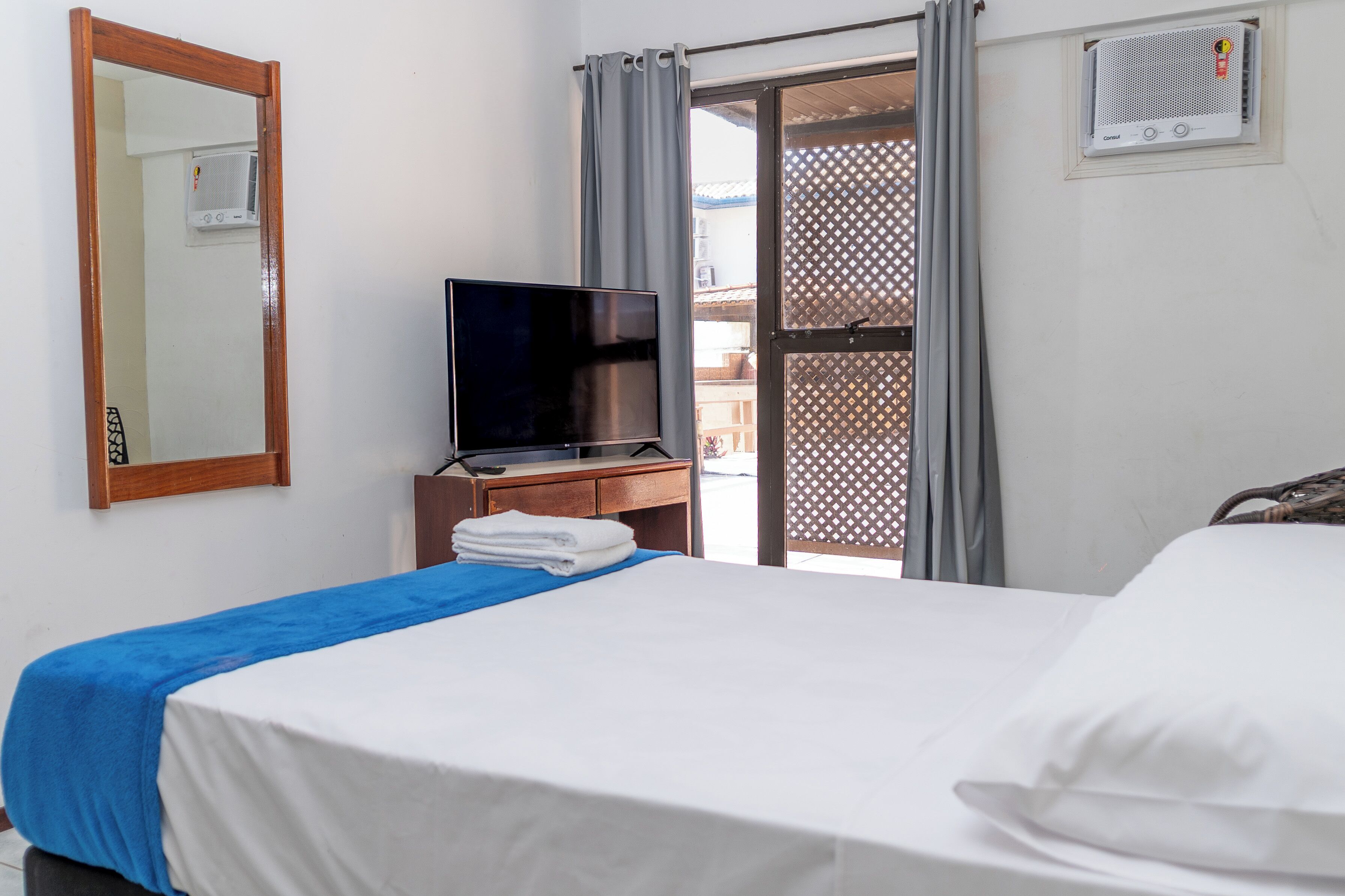 A hotel room with a bed, a TV on a wooden stand, a window with curtains, and a balcony with a lattice design.