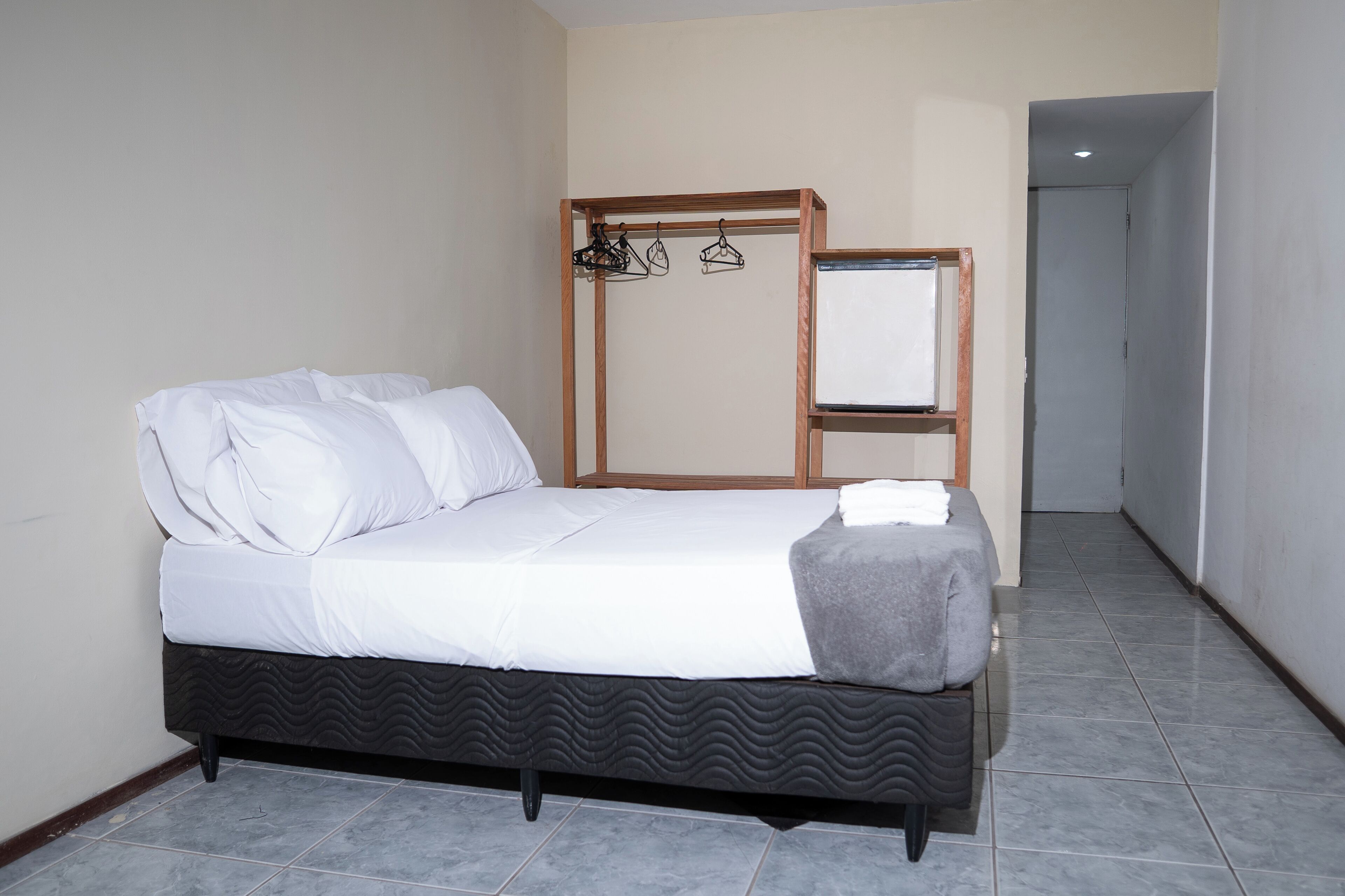 A neatly made bed with white linens, a dark patterned bed base, and a grey throw blanket. There is a wooden shelf with clothes hanging on it and a mirror on the wall.