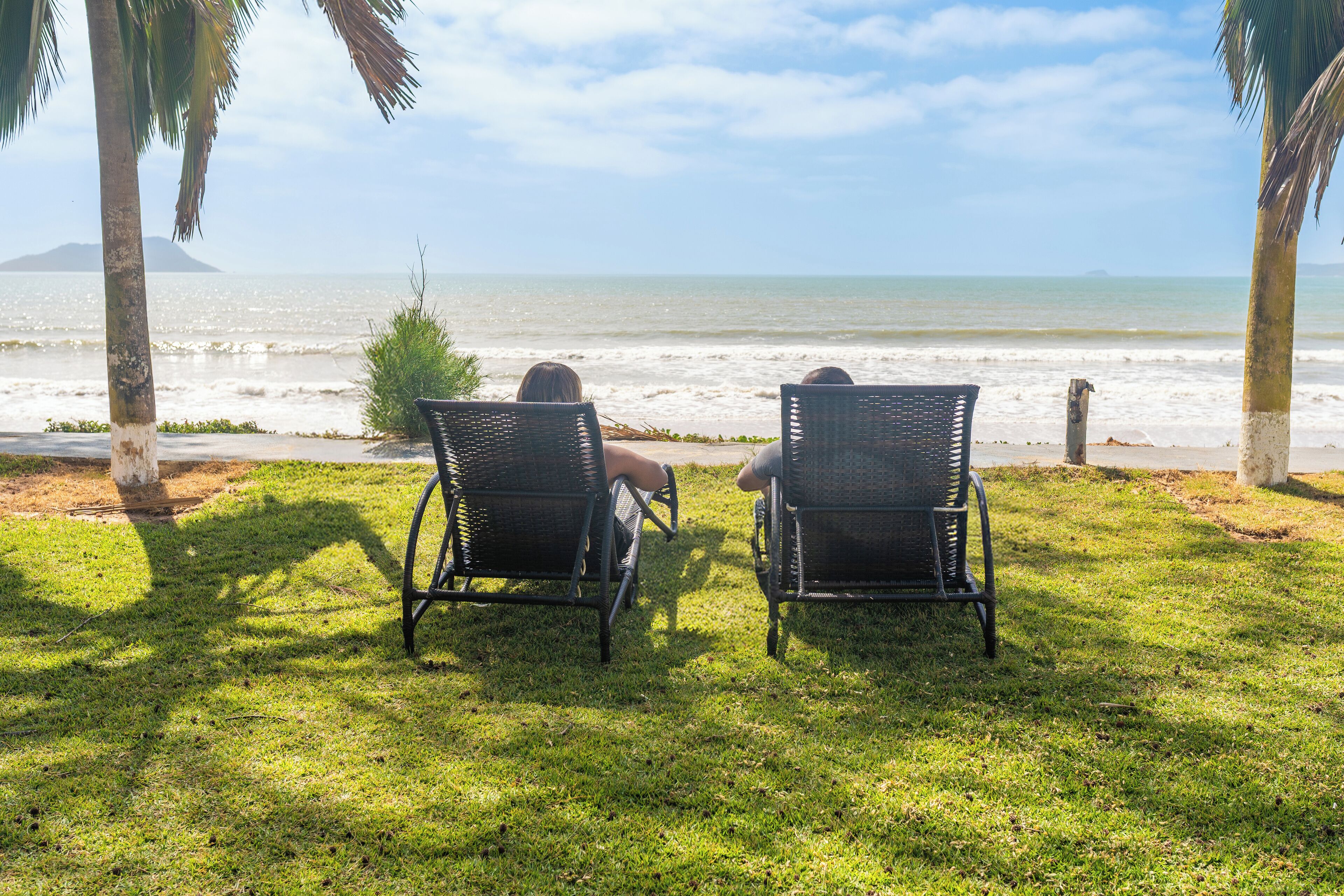 Two wicker chairs facing the ocean with a view of the beach and palm trees.
