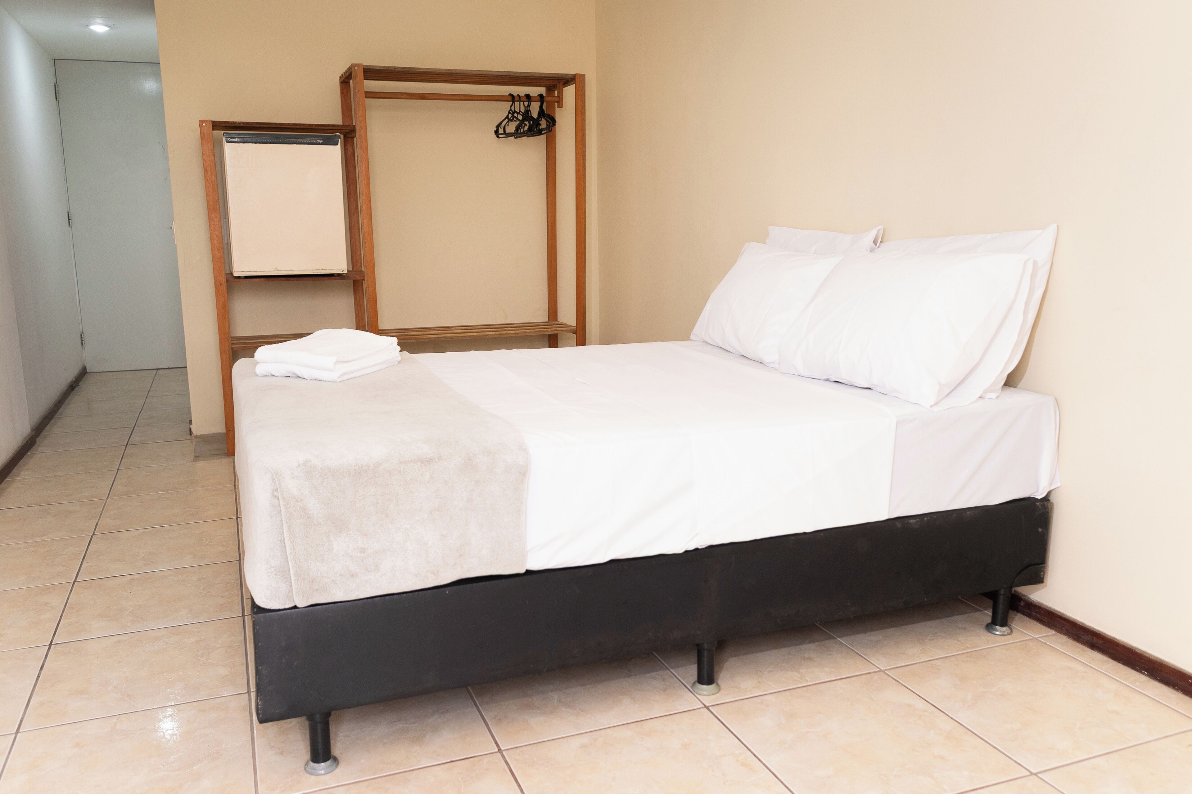 A neatly made bed with white linens, a wooden headboard, and a small shelf with a mirror and hanging clothes.