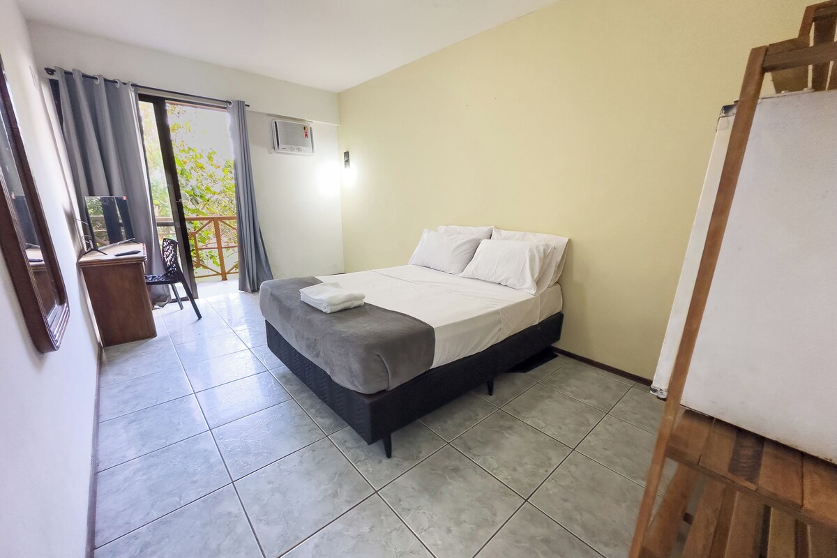 Une chambre à coucher avec un lit, un bureau et une chaise. Il y a une fenêtre avec des rideaux, et de la fenêtre, on peut voir la verdure à l’extérieur.