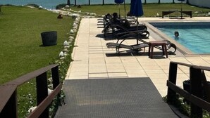 Zona de piscina con sillas de descanso y sombrillas, una palmera y vista al océano.
