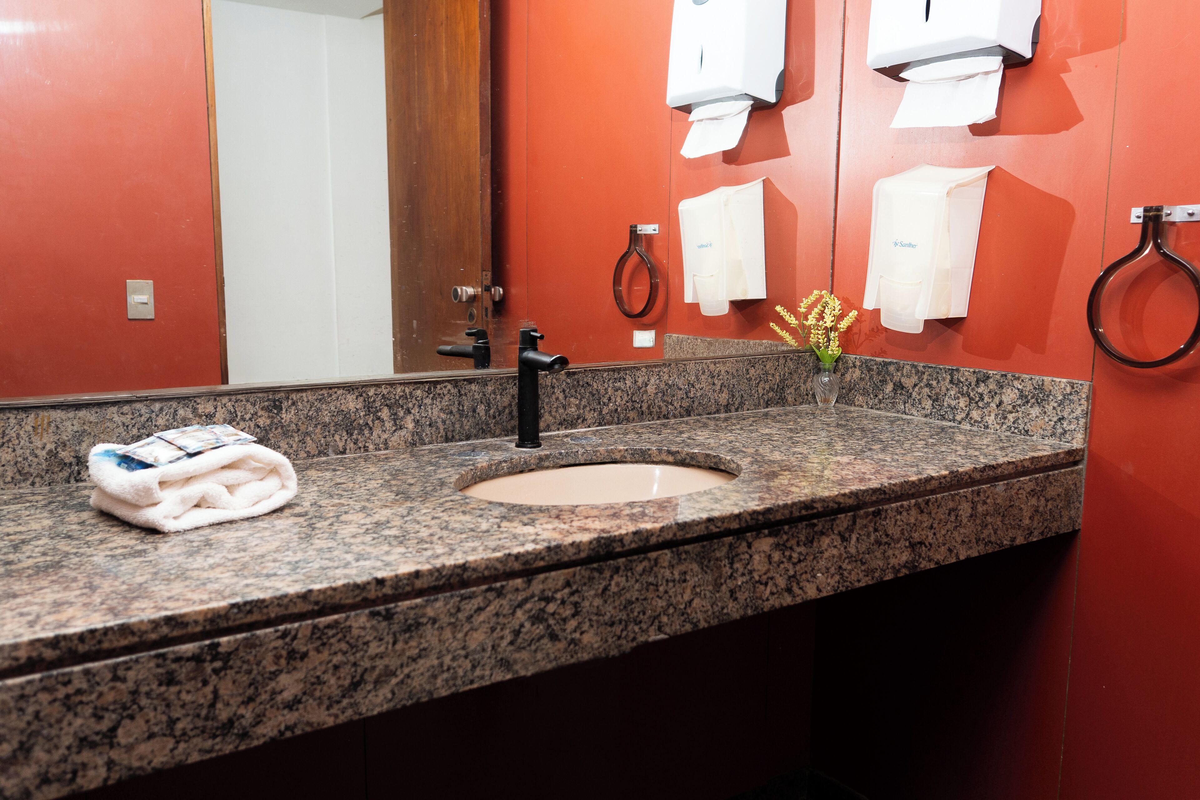 A bathroom with a granite countertop, a sink, a mirror, a towel dispenser, and a toilet paper holder.