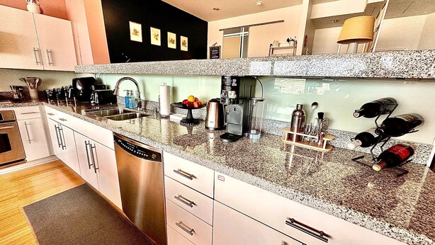 A modern kitchen with white cabinets, granite countertops, a dishwasher, a coffee maker, and wine bottles on a rack.