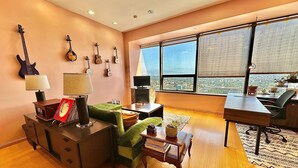 A modern living room with a wooden floor, a green sofa, a wooden coffee table, and a desk with a chair. There are guitars on the wall and a large window with a view of the city.