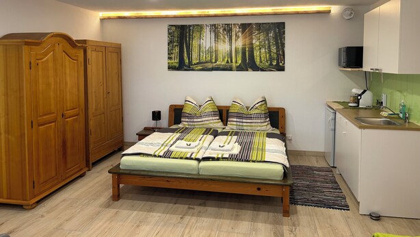 A bedroom with a wooden bed, a painting of a forest, a kitchenette with a microwave, and a wardrobe. - Wandlhof vacation apartment (Niederösterreich)