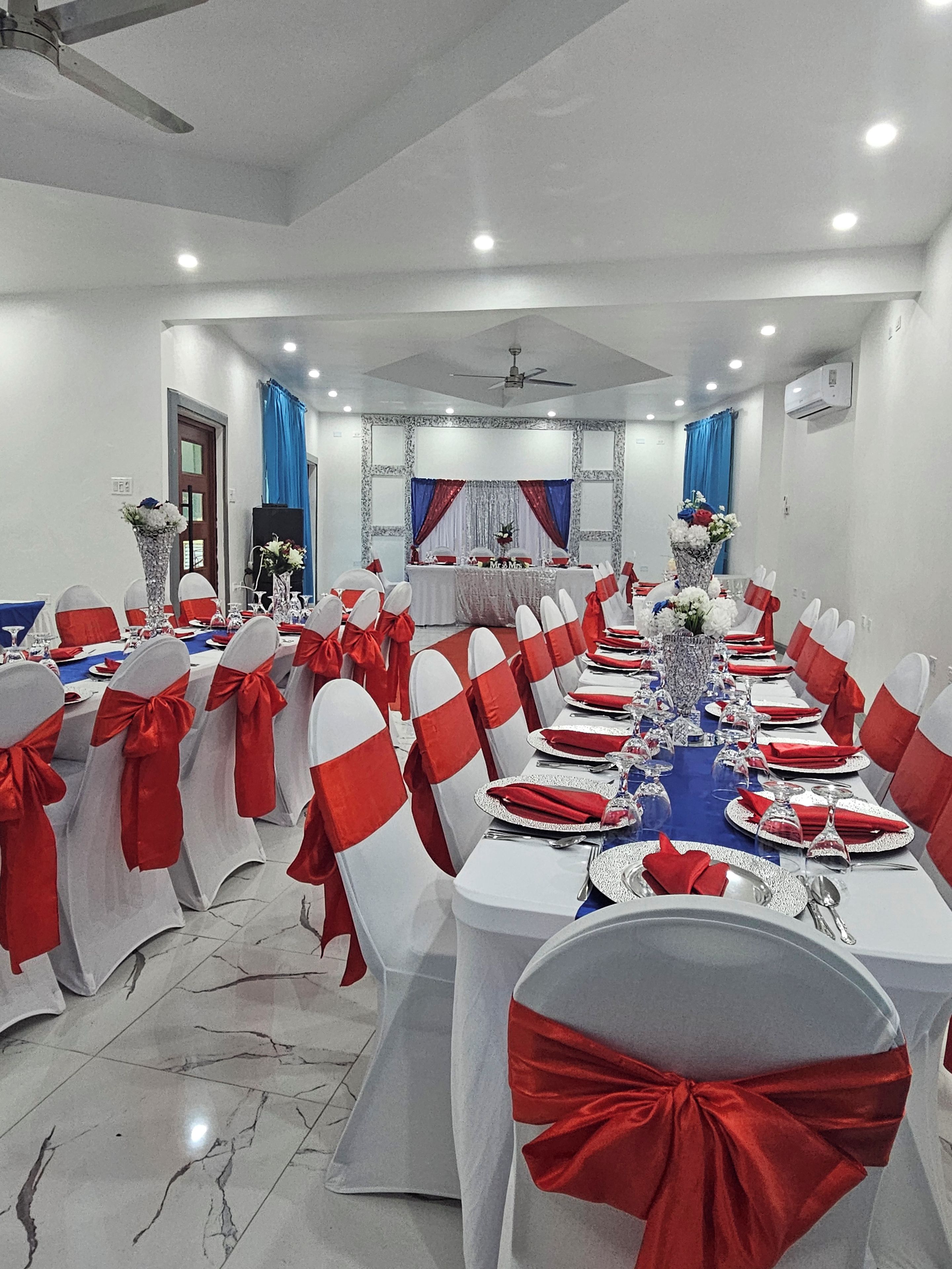 A banquet hall with round tables, white chairs with red sashes, and a blue tablecloth.