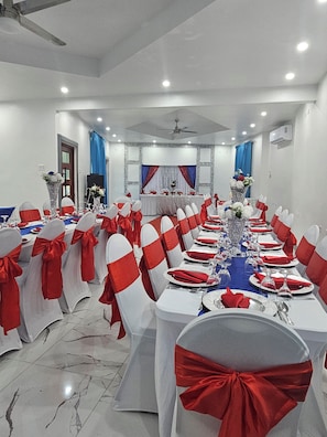 A banquet hall with round tables, white chairs with red sashes, and a blue tablecloth.