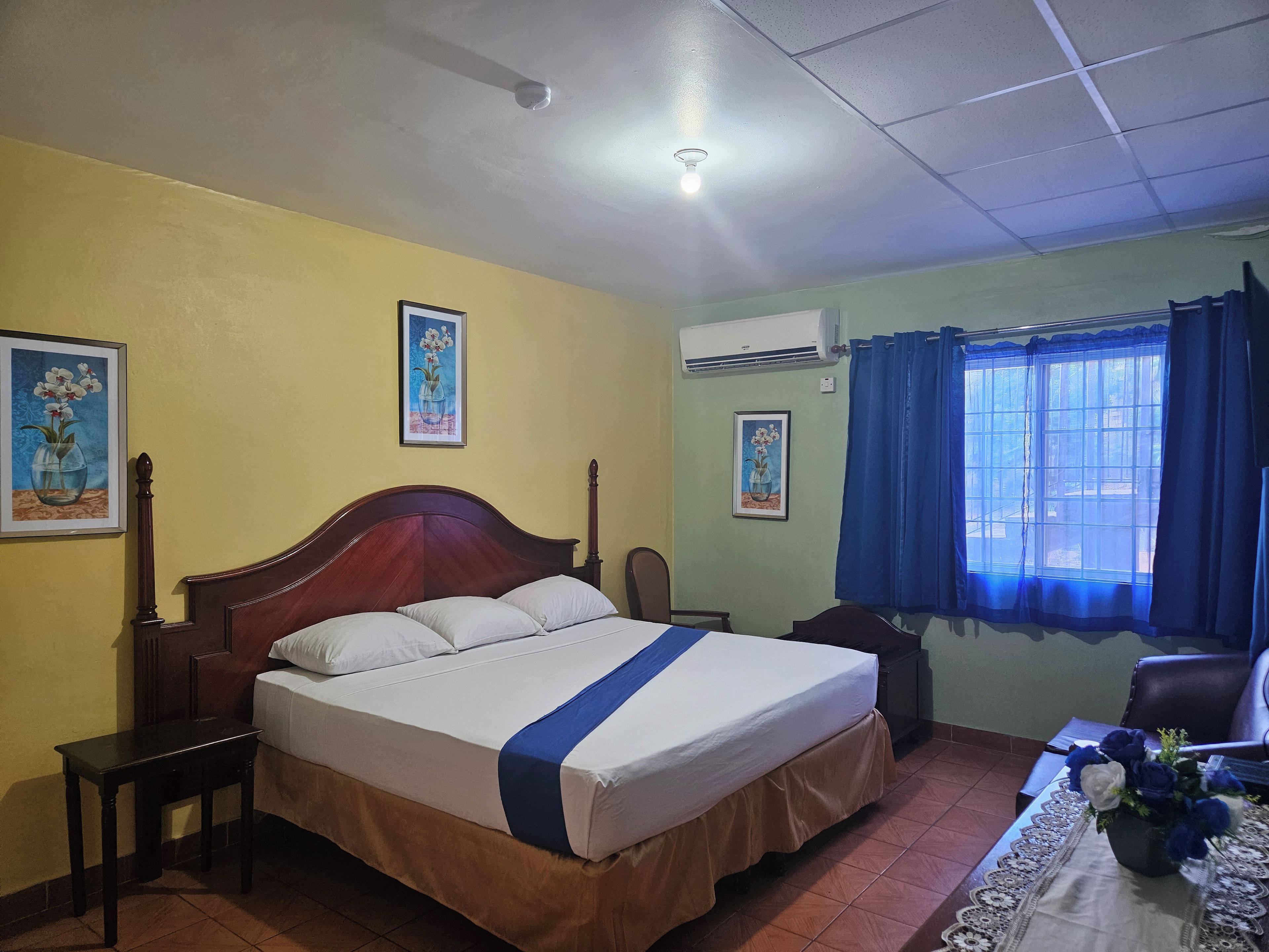 Un dormitorio con una cama, una mesita de noche, una silla, una mesa con un jarrón de flores y dos unidades de aire acondicionado fijadas a la pared.