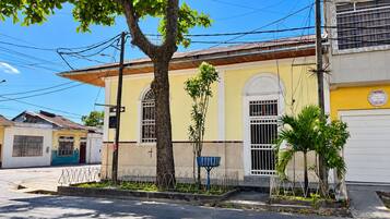 A building with a yellow facade, a large tree in front, and a metal fence.