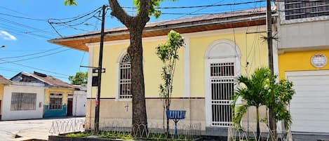 A building with a yellow facade, a large tree in front, and a metal fence.