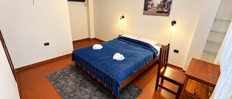 A hotel room with a bed, a desk, a chair, a rug, a picture on the wall, and a ceiling-mounted device.