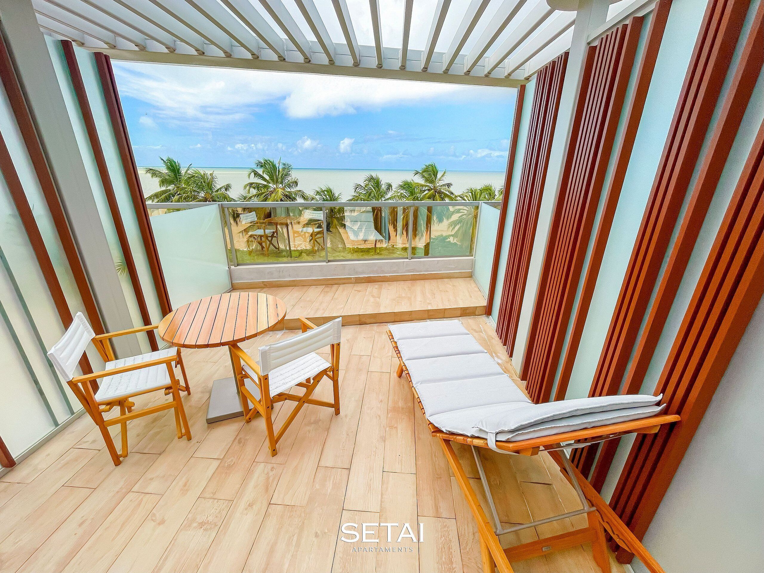 A balcony with a wooden floor, two wooden chairs, a small round table, and a white cushioned lounge chair.