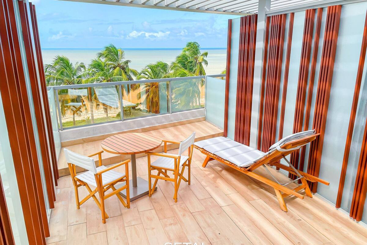 A balcony with a wooden table and chairs, a lounge chair, and a view of the beach.