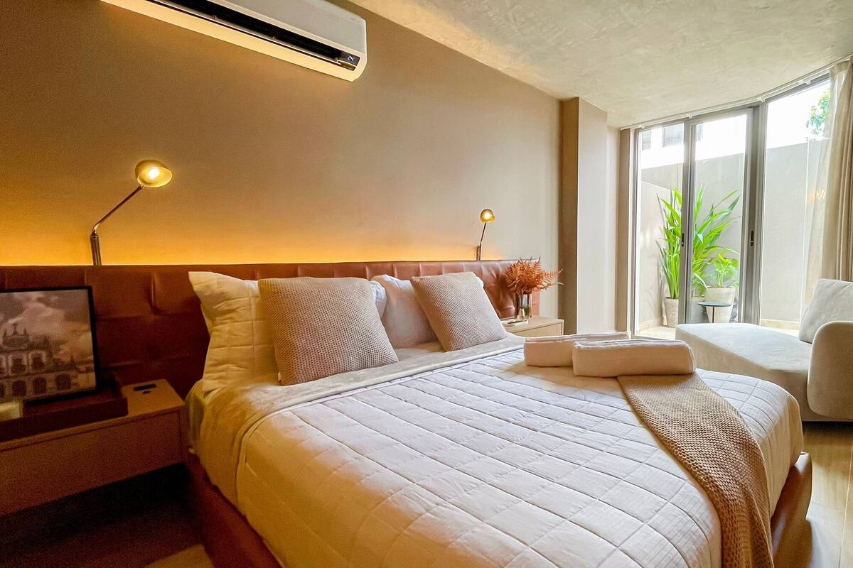 A modern hotel room with a large bed, bedside lamps, a nightstand, a small table with a plant, and a view of greenery through the window.