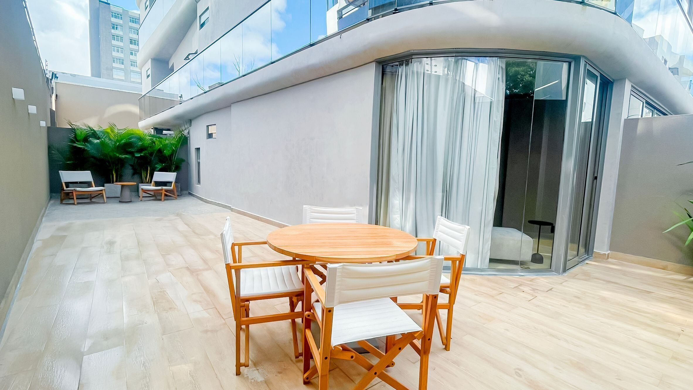 Un patio exterior moderno con una mesa redonda de madera y sillas blancas acolchadas, rodeado de plantas en macetas y un ventanal de cristal que da a un balcón.
