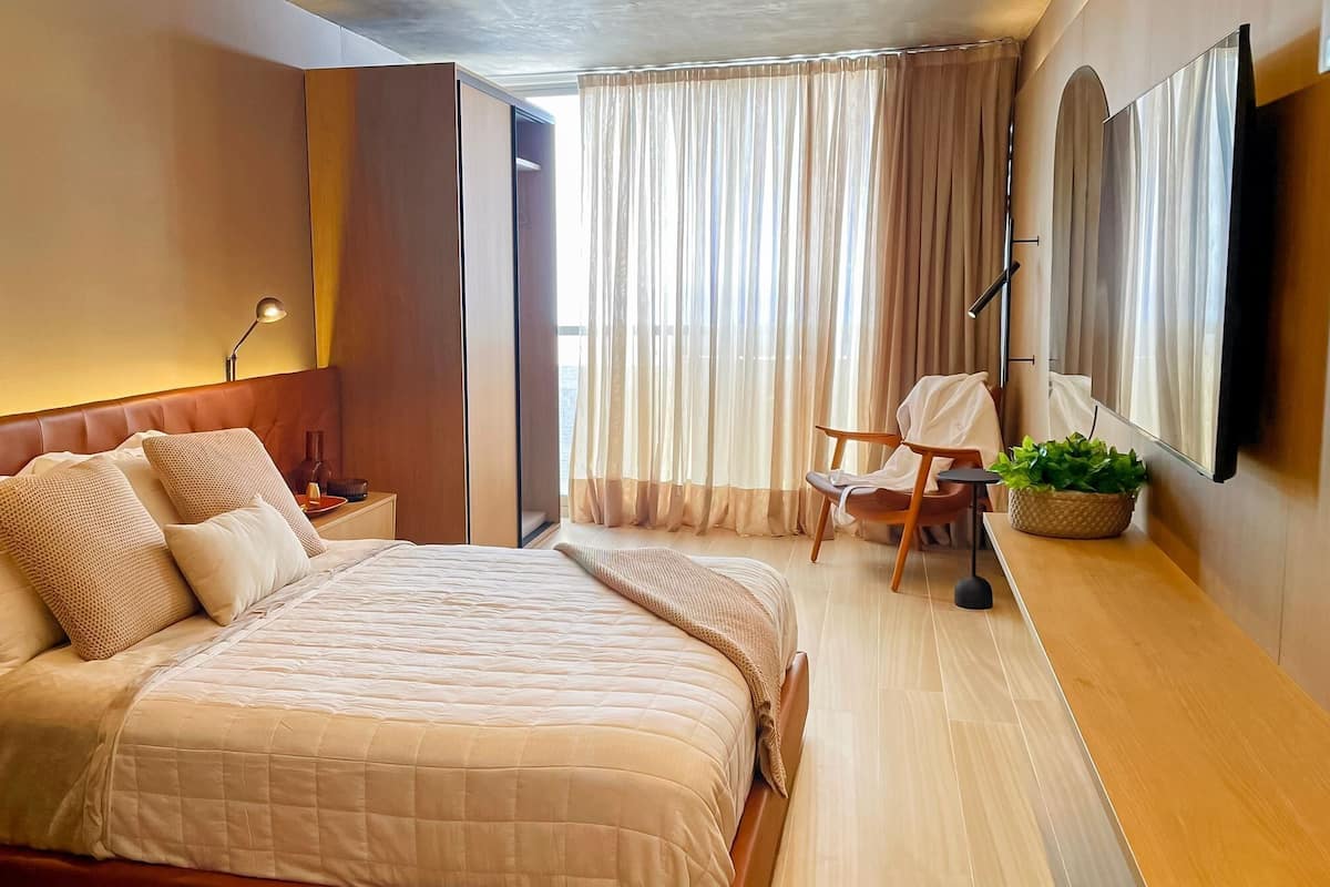 A hotel room with a large bed, a chair, a small table, a potted plant, and a mirror.