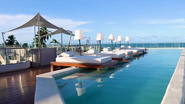 A poolside area with lounge chairs and umbrellas, a clear view of the ocean, and a wooden deck. - Yacht - by Setai Apartaments (João Pessoa)