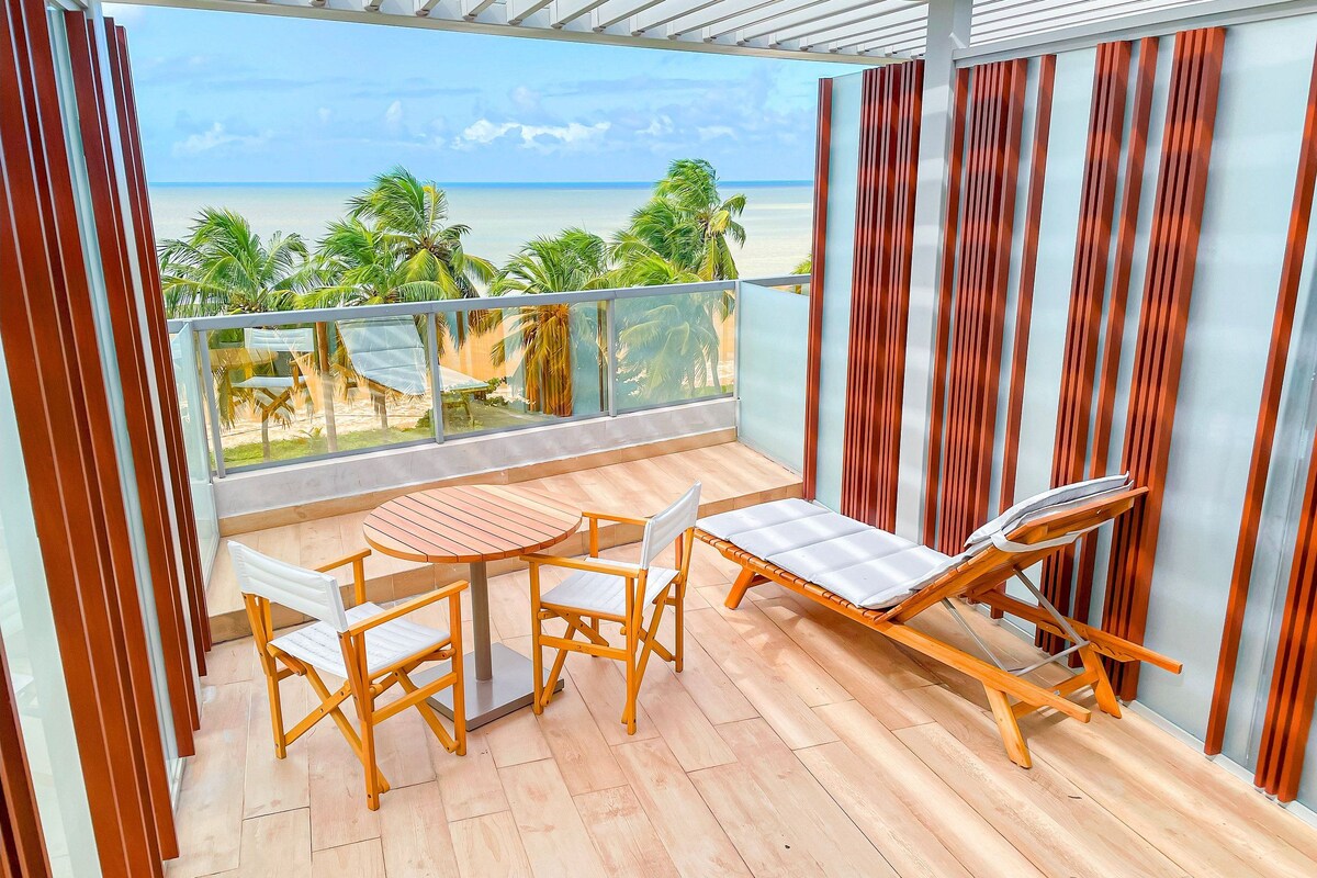 Un balcon avec une table et des chaises en bois, un fauteuil de relaxation et une vue sur la plage.