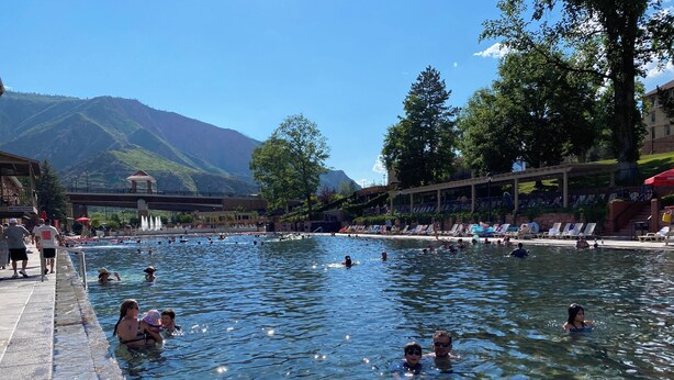 Una piscina con gente nadando y tomando el sol, rodeada de árboles y una montaña al fondo.