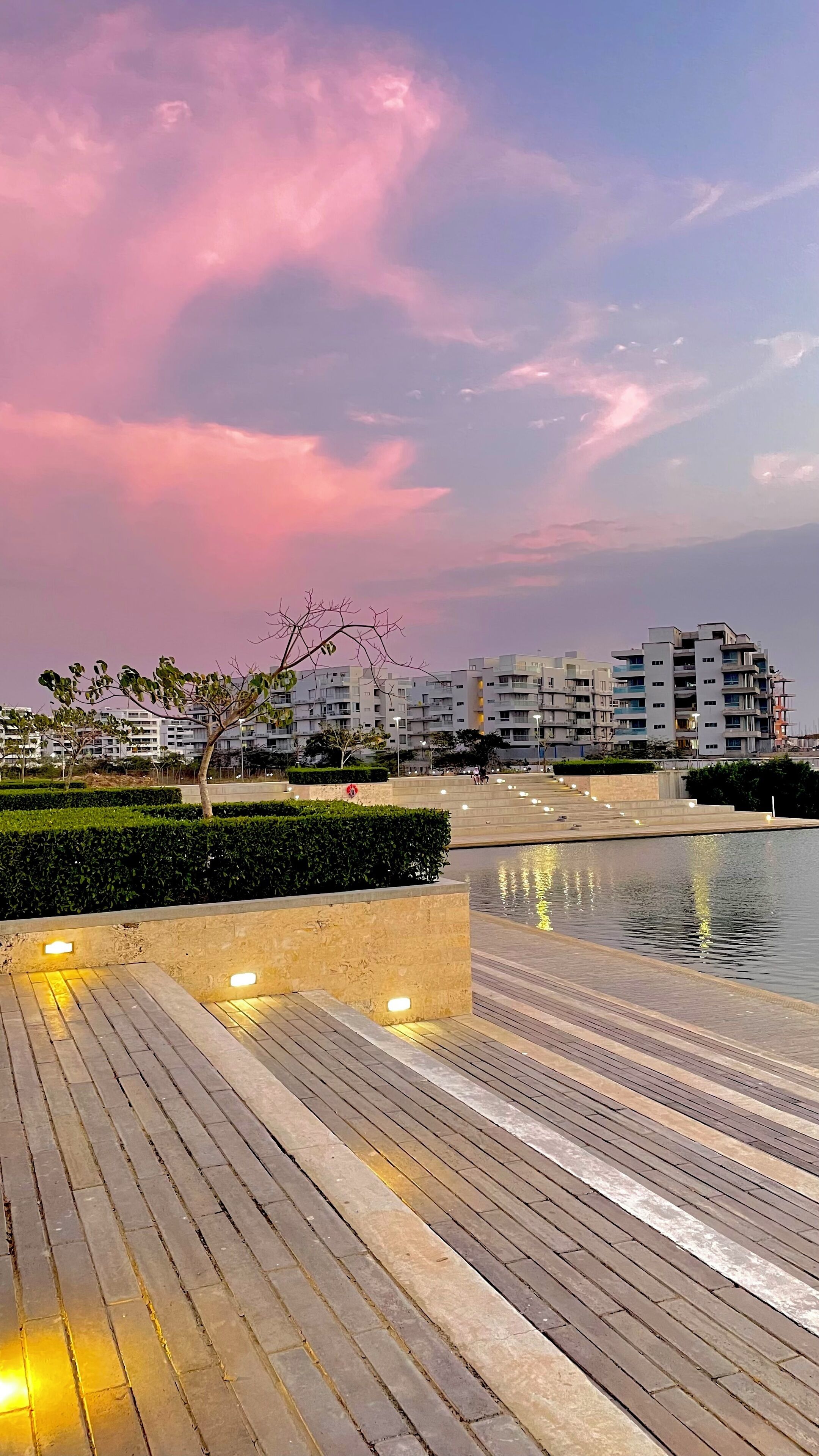 A waterfront promenade with a wooden deck, a water body reflecting lights, and modern residential buildings in the background.