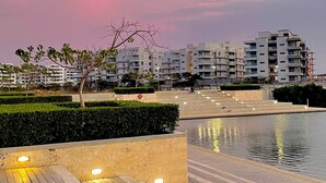 En strandpromenad med ett trädäck, en vattenyta som speglar ljusen och moderna bostadshus i bakgrunden.